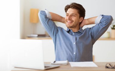 Gülümseyen bir adam önünde dizüstü bilgisayarla masada oturuyor, kolları başının arkasında rahatlıyor..