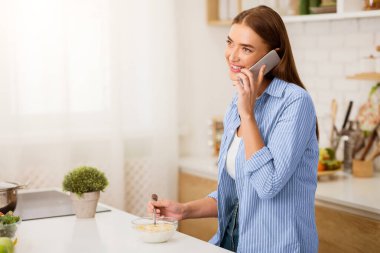 Mavi çizgili gömlekli bir kadın mutfakta kahvaltı ederken telefonla konuşuyor..
