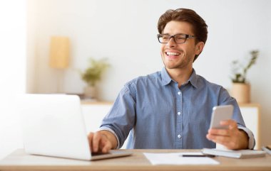 Gözlüklü bir adam bir masada oturup dizüstü bilgisayarı üzerinde çalışır, diğer elinde bir telefon tutarken gülümser..