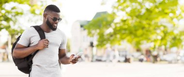 Güneş gözlüğü ve sırt çantası takan bir adam akıllı telefonuna bakarken sokakta yürüyor..