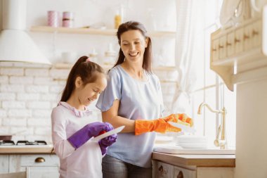 Bu resim modern bir mutfakta bulaşıkları yıkayan bir anne ve kızı gösteriyor. Annem turuncu eldiven giyiyor ve lavaboda bir tabak yıkıyor. Kızı mor eldiven giyiyor ve elinde bir tabak tutuyor.