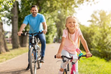 Genç bir kız, parktaki asfalt bir yolda pembe bir bisiklet sürerken gülümsüyor. Babası onun arkasında, o da bisiklet sürüyor. İkisi de açık havada güneşli bir günün tadını çıkarıyor..