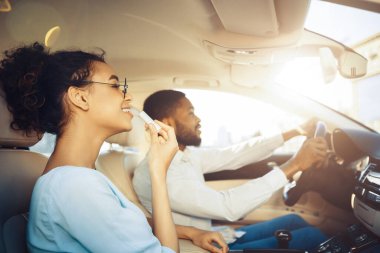 Kıvırcık saçlı genç siyah bir kadın bir arabanın yolcu koltuğunda otururken dudak kremi sürüyor. Arabayı bir adam kullanıyor. Kadın gülümsüyor ve dikiz aynasında yansımasına bakıyor..