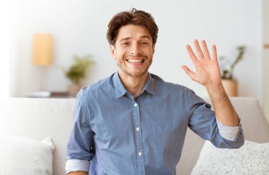 Oturma odasındaki kanepede oturan bir adam gülümseyip elini sallıyor..