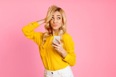 Uzun sarı saçlı genç bir kadın akıllı telefon tutarken sarı bir gömlek ve beyaz bir şort giyiyor. O, şaşkın bir ifadeyle yukarı bakıyor. Arka plan pembe..