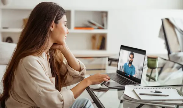 stock image A young woman sits at a desk in her home office, attentively listening to a video conference on her laptop. The room is well-lit, and the woman is dressed casually in a light-colored shirt.