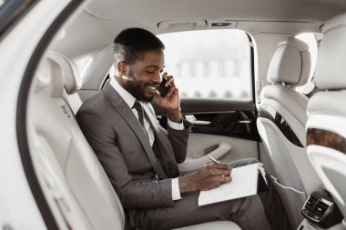 Takım elbiseli Afrikalı Amerikalı işadamı lüks bir arabanın arka koltuğunda oturur ve telefonuyla konuşurken not defterine not alır. Odaklanmış ve profesyonel görünüyor..