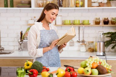 Mutfakta önlük giymiş ve yemek kitabı okuyan bir kadın var. Tezgahtaki taze ürünlerin de gösterdiği gibi yemek hazırlıyor..