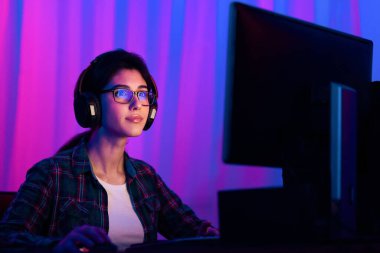 Kulaklık takan genç bir kadın bilgisayarın başında oturuyor, neon ışıklarla aydınlatılıyor. O, yüzünde ciddi bir ifadeyle oyununa odaklanmış. O rekabetçi bir oyun oynuyor gibi görünüyor.