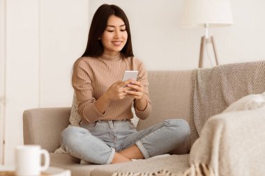 Kanepede bacakları çapraz oturan bir kadın elindeki akıllı telefona bakıyor. Kahverengi bir kazak ve kot pantolon giyiyor..