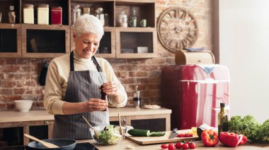 Son sınıftan bir kadın mutfağında salata hazırlıyor. Elinde bir biber değirmeni tutuyor, yemeğine biraz baharat katmaya hazır. Mutfak sıcak ve davetkar.
