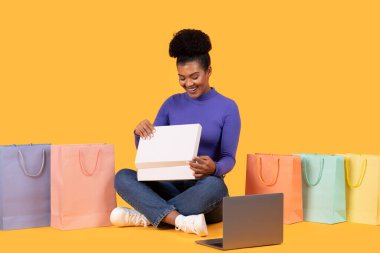 Mutlu genç bir kadın sarı arka planda oturuyor, alışveriş torbalarıyla çevrili. Beyaz bir kutu açıyor, içeriği konusunda heyecanlı görünüyor. Önünde bir dizüstü bilgisayar duruyor.