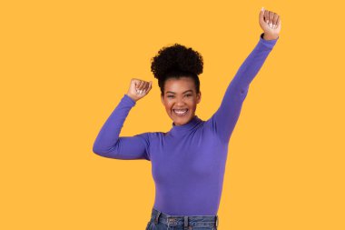 Koyu kahverengi tenli ve büyük afro saçlı genç bir kadın uzun kollu mor bir gömlek giyiyor. Kolları havada sarı bir arka planın önünde duruyor ve yüzünde kocaman bir gülümseme var..