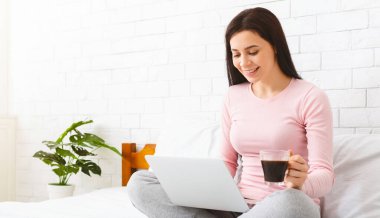 Bir kadın bir elinde bir fincan kahveyle yatakta oturuyor ve dikkatle bilgisayar ekranına bakıyor. Laptopta görünen içeriğe odaklanmış ve meşgul görünüyor, boş bir alan.