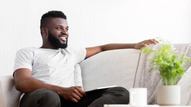 Genç bir Afrikalı Amerikalı adam kanepede oturuyor ve gülümsüyor. O, beyaz bir tişört ve siyah pantolon giyiyor ve eli bir tabletin üzerinde. Sağında bir bitki var.
