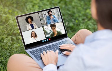 Bir kadın öğrenci çimlerin üzerinde oturuyor ve video konferans toplantısı için dizüstü bilgisayar kullanıyor. Ekranda her bir video fayansında dört kişi gösteriliyor