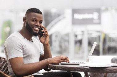 Bir adam dışarıda bir masada oturuyor, telefonuyla konuşurken dizüstü bilgisayarıyla çalışıyor. Gülümsüyor ve sohbet ediyor gibi görünüyor..