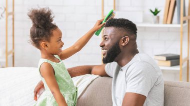 Kıvırcık saçlı genç bir siyah kız babasının saçını yeşil bir fırçayla tararken o bir kanepede oturuyor. Kız gülümsüyor ve babasına bakıyor, o ise gülümsüyor ve ona bakıyor..