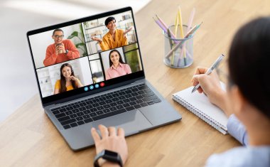 Kadın dizüstü bilgisayardaki bir video konferansına katılırken not defterine not alıyor. Ekranda dört kişi ayrı video çerçevelerinde görüntüleniyor
