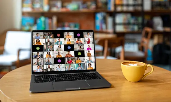 stock image A laptop computer sits on a wooden table in a cafe. The laptop screen displays a video conference meeting with multiple participants from different backgrounds.