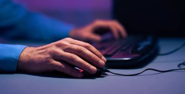 Görüntü, bilgisayar faresi kullanan bir elin yakın çekimini gösteriyor. Kişi dizüstü bilgisayarın önünde oturuyor ve odadaki tek ışık ekrandan geliyor.