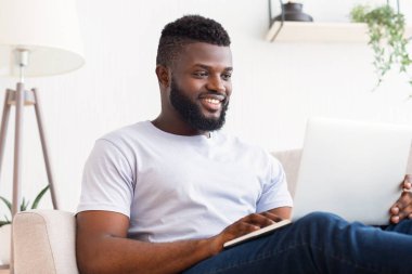Sakallı Afrikalı Amerikalı genç bir adam oturma odasında bir kanepede oturuyor ve bir dizüstü bilgisayar kullanıyor. Gülümsüyor ve mutlu görünüyor..