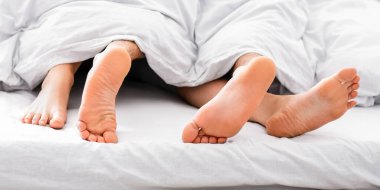 Bu resim, beyaz yatağın altından sarkan iki çıplak ayak görüntüsünü gösteriyor. Ayaklar iki kişiye ait gibi görünüyor. Muhtemelen bir çifte. Yatakta birlikte yatıyorlar.