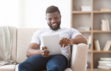 Afro-Amerikalı bir adam oturma odasındaki beyaz bir kanepede oturuyor, tablet kullanırken gülümsüyor. Beyaz bir tişört ve kot pantolon giyiyor. Kanepe de kitaplığın yanında..