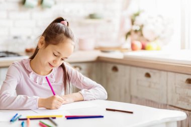 Pembe tişörtlü, uzun kahverengi saçlı genç bir kız mutfak masasında oturuyor, bir kağıt parçasına renkli kalemlerle çizim yapıyor. Mutfak parlak ve havadar, arka planda bir pencere var..