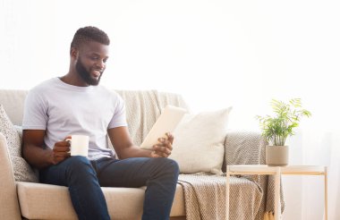 Sakallı genç siyah bir adam parlak bir oturma odasında bej bir kanepede beyaz bir tablet ve elinde bir fincan kahve ile oturuyor. Tablete bakıyor, gülümsüyor.