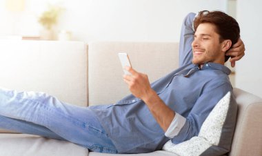 Bir adam akıllı telefonunu kullanarak kanepede dinleniyor. Gülümsüyor ve eğleniyor gibi görünüyor..