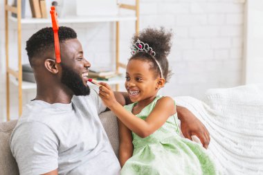 Prenses tacı takan genç bir kız beyaz bir kanepede birlikte otururken babasının dudaklarına ruj sürüyor. Kızın saçı afro ve yeşil bir elbise giyiyor..