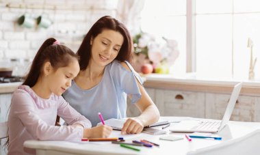 Genç bir kız mutfak masasında oturuyor, renkli kalemlerle çizim yapıyor. Annesi yanında oturuyor, ona rehberlik ediyor ve gülümsüyor. Mutfak aydınlık ve havadar, doğal ışık sağlayan bir pencere var..