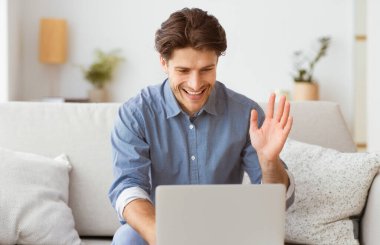 Bir adam koltukta oturuyor, gülümsüyor ve önünde diz üstü bilgisayara el sallıyor..
