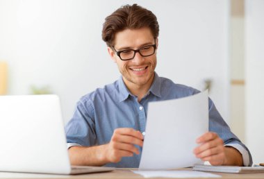 Bir adam elinde dizüstü bilgisayarla masada otururken belgeyi okurken gülümsüyor..