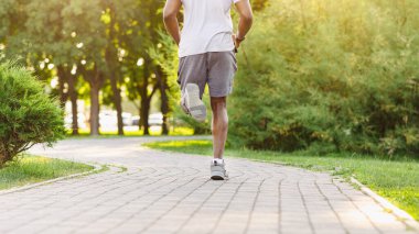 Atletik giysili bir adam yeşil çimenler ve ağaçlarla çevrili bir parkta asfalt bir yolda koşuyor..