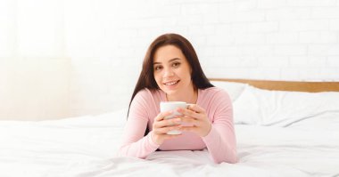 Bir kadın elinde bardakla yatakta uzanıyor. Yumuşak çarşaflar ve yastıklar rahat bir zemin sağlıyor. İçeceğiyle bir dakikalık rahatlığın tadını çıkarıyor..