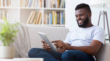 Gülümseyen siyah bir adam oturma odasında beyaz bir kanepede oturur. Rastgele giyiniyor ve bir tablet bilgisayar kullanıyor, rahat görünüyor ve boş zamanlarının tadını çıkarıyor. Oda iyi aydınlatılmış ve dekore edilmiş.