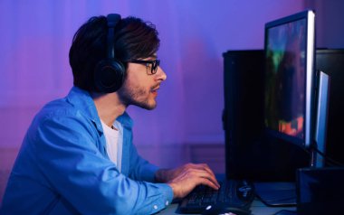 Adamın biri bilgisayarın önünde kulaklıkla oturmuş video oyunu oynuyor. Ekrana odaklanmış durumda ve elleri klavyede. Oda mor ışıkla aydınlatılmış.