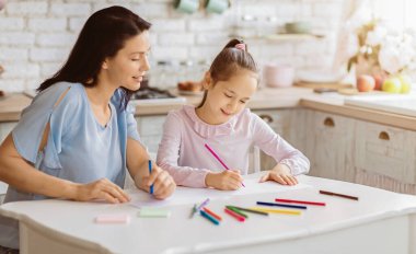 Bir anne ve kızı mutfak masasında oturuyor, ikisi de çizim yapıyor. Kız dikkatlice çizimine odaklanırken anne izliyor ve gülümsüyor. Masa renkli kalemlerle kaplı.