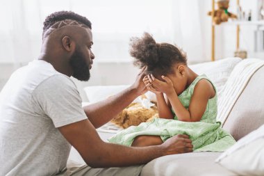 Bir baba kanepeye oturur ve üzgün kızını teselli eder. O, elleriyle yüzünü örterken elini onun başına koyar. O beyaz bir tişört giyiyor ve o yeşil bir elbise giyiyor..