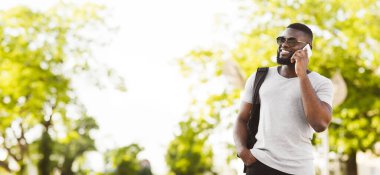 Sırt çantalı genç bir adam dışarıda telefonla konuşurken gülümsüyor. Yeşil ağaçların bulanık arka planının önünde duruyor..