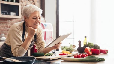 Önlüklü yaşlı bir kadın mutfağında tarif bulmak için tablet kullanıyor. Tezgah salatalık, domates, dolma biber ve brokoli gibi taze ürünlerle dolu..