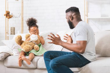 Afro-Amerikalı bir baba kanepeye oturur, gülümser ve elleriyle gözlerini kapatır. Genç kızı yanında oturuyor, elleriyle gözlerini kapatıyor, oyuncak ayıyı tutuyor.