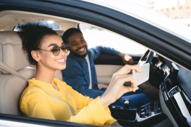 Afrikalı Amerikalı genç bir çift arabada oturup selfie çekiyor. Kadın güneş gözlüğü ve sarı bir gömlek giyiyor ve adam mavi bir gömlek giyiyor. İkisi de gülümsüyor ve mutlu görünüyorlar.