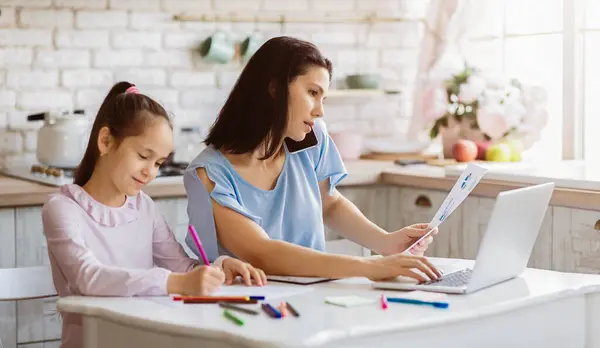 stock image A mother is working on her laptop while her daughter colors at a table in the kitchen. The mother is on the phone and appears to be busy with work. The daughter is focused on her coloring