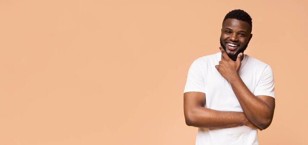 A young African American man in a white t-shirt smiles brightly while standing against a peach colored background. He has a thoughtful expression and his right hand is resting on his chin, copy space