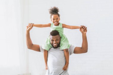 Neşeli bir siyah baba, genç kızına evin içinde bir gezinti yaptırıyor. Kız yeşil bir elbise giyiyor ve kollarını açmış, baba gülümsüyor ve kameraya bakıyor..