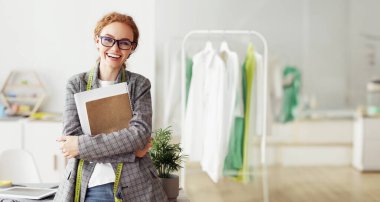 Mutlu bir kadın, bir moda tasarımcısı, stüdyosunda duruyor, elinde bir defter, ve güvenle gülümsüyor..
