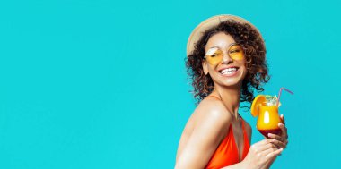 Güneş gözlüğü ve hasır şapka takan Afro-Amerikalı genç bir kadın elinde parlak mavi bir arka planda tropik bir kokteyl tutarken gülümsüyor..